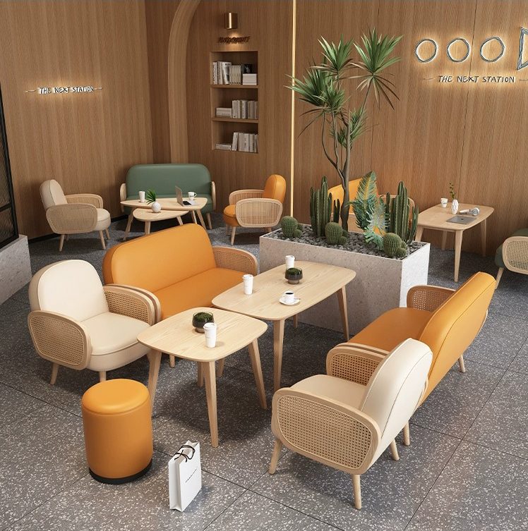 A modern cafe lounge with rattan armchairs, orange and beige sofas, and wooden tables, complemented by cactus decor and a minimalist bookshelf in the background.