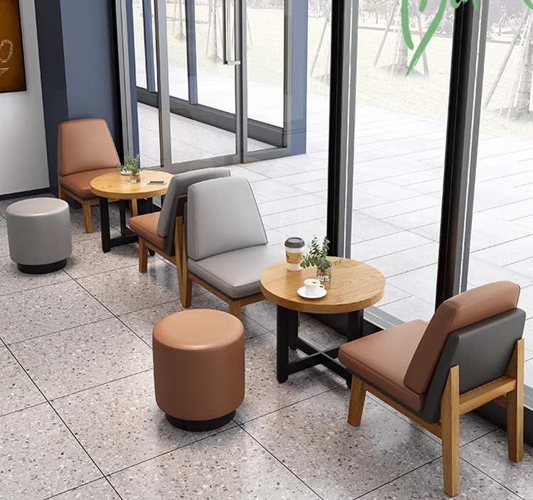 A modern cafe seating area featuring leather chairs with wooden frames and round tables. The space includes gray, brown, and beige leather stools and wooden tables near large windows.