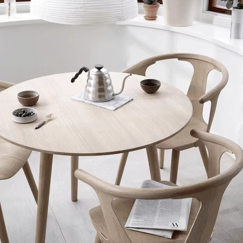 Minimalist dining area featuring light wooden chairs, a round wooden table, and large windows for natural lighting.