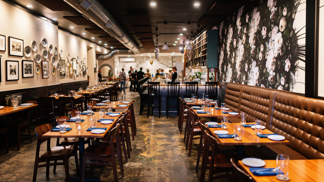 Modern restaurant interior with floral-themed wallpaper, wooden tables and chairs, and a cozy bar area.