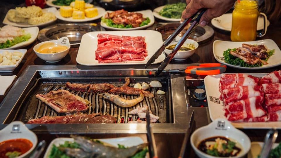 Hot pot grill with fresh meat slices, seafood, vegetables, and dipping sauces in a restaurant setting.