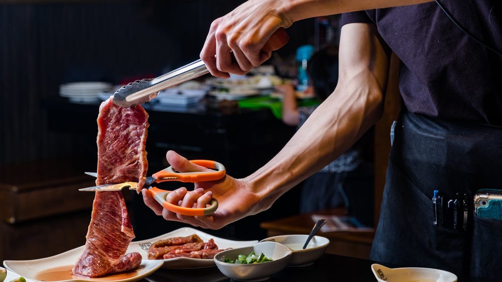 Chef slicing raw meat with tongs and scissors in a restaurant kitchen, preparing for grilling or serving.