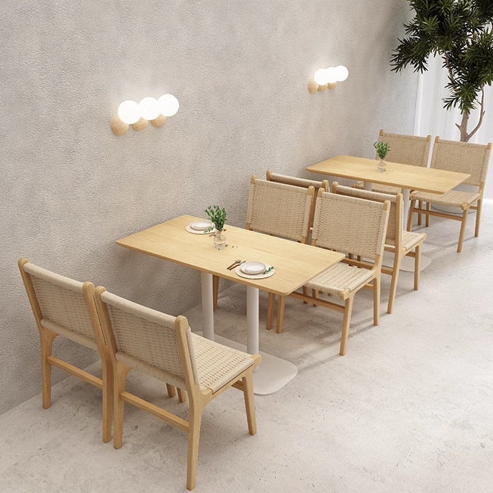 Minimalist dining area featuring light wooden tables, woven chairs, and simple table settings with decorative plants.