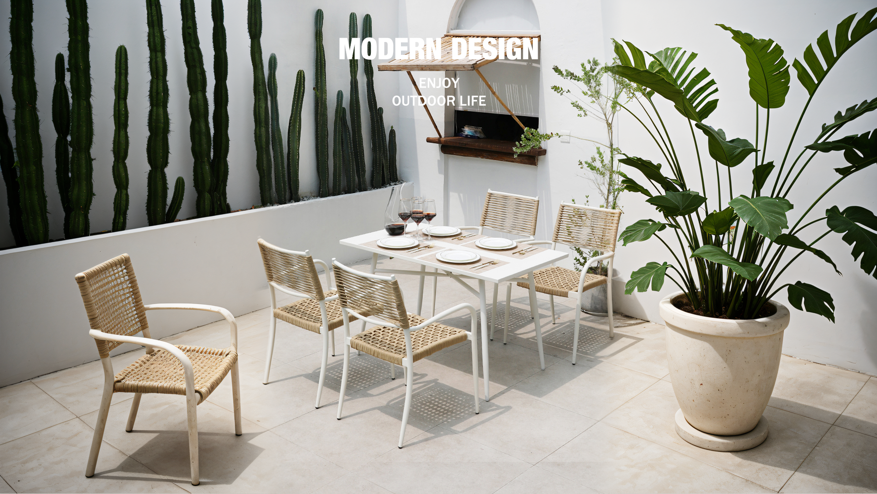 Modern outdoor dining set with rattan chairs and a white table, surrounded by cactus and large tropical plants.