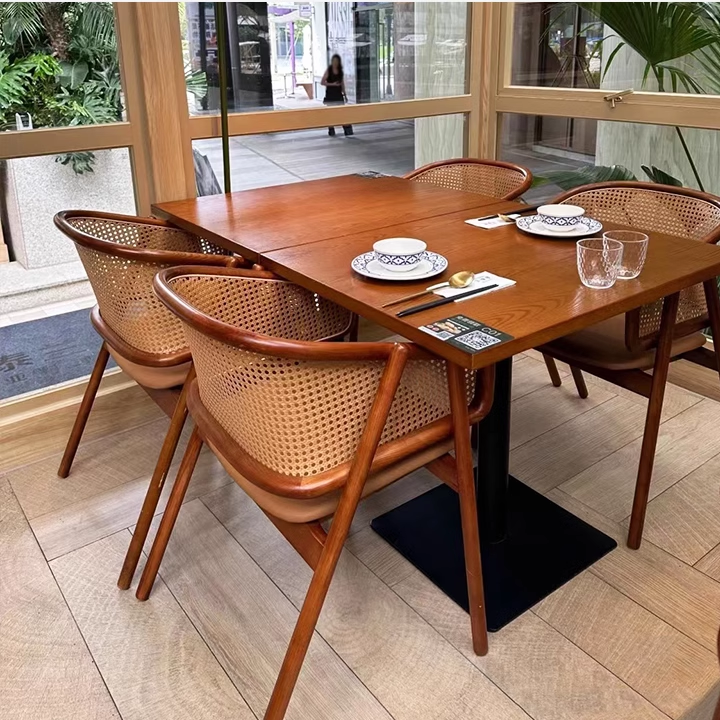 Restaurant interior featuring a wooden dining table with cane-back chairs, set with plates, glasses, and chopsticks, against a bright window view.