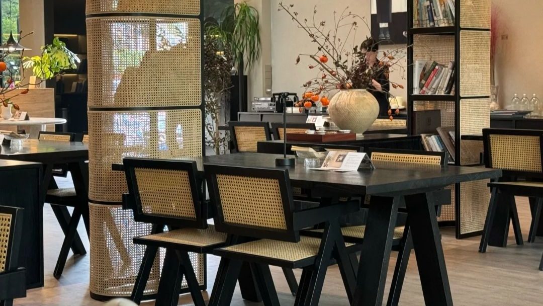 A modern woven shelving unit displayed in a cafe, featuring bamboo caning, black metal frames, and books on the shelves. Nearby tables with black chairs complete the decor.