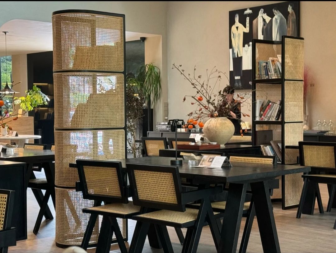 A modern woven shelving unit displayed in a cafe, featuring bamboo caning, black metal frames, and books on the shelves. Nearby tables with black chairs complete the decor.